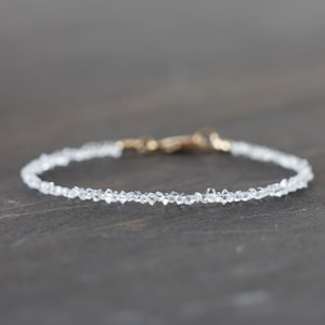 May include: Delicate bracelet featuring small, clear, faceted beads strung together. The bracelet has a gold-colored clasp. The beads appear to be evenly spaced, creating a subtle sparkle. The jewelry is displayed on a dark, neutral surface.