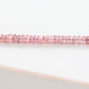 Op de afbeelding: Een streng kleine, gefacetteerde roze edelstenen. De edelstenen hebben een lichte, doorschijnende roze kleur en zijn in een lijn geregen. De achtergrond is een effen wit oppervlak.