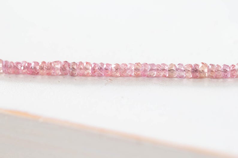 Op de afbeelding: Een streng kleine, gefacetteerde roze edelstenen. De edelstenen hebben een lichte, doorschijnende roze kleur en zijn in een lijn geregen. De achtergrond is een effen wit oppervlak.