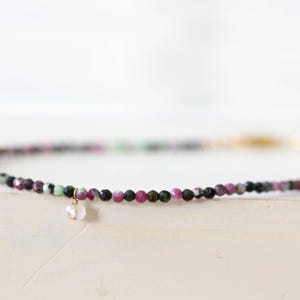 May include: Delicate beaded necklace featuring small, colorful gemstones in shades of pink, green, and black. A small, clear crystal pendant hangs from the necklace. The necklace is set against a soft, neutral background.
