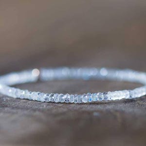 May include: A delicate bracelet made of small, clear, faceted beads. The beads are strung together, forming a circular shape, resting on a neutral surface. The bracelet has a subtle sparkle, possibly made of a gemstone or crystal.