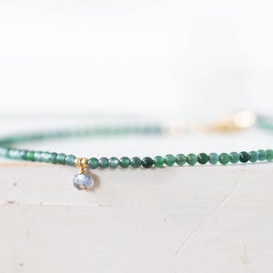 May include: Delicate green beaded bracelet with a small, iridescent charm. The bracelet features small, round green beads strung together with a gold-colored clasp. The charm is a light blue color. The bracelet is displayed on a white surface.