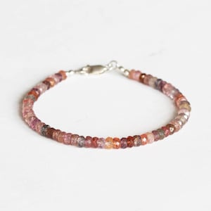 May include: A delicate bracelet featuring small, faceted beads in shades of pink, red, and brown. The beads are strung together with a silver clasp. The bracelet is displayed on a white background.