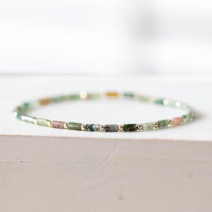 May include: A delicate bracelet featuring small, rectangular beads in shades of green, pink, and gold. The beads are strung together with gold-colored accents. The bracelet is displayed on a white surface, showcasing its minimalist design.