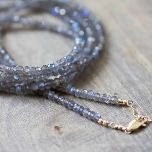 May include: A multi-strand necklace composed of small, faceted gray beads with blue and iridescent flashes. The necklace has a gold-colored clasp and chain, resting on a wooden surface. The beads are strung together to create a layered effect.