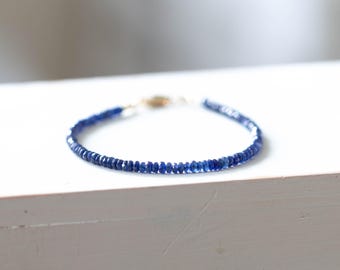 Puede incluir: Una delicada pulsera con pequeñas piedras preciosas azules facetadas ensartadas. La pulsera tiene un cierre dorado y se muestra sobre una superficie blanca. Las piedras preciosas parecen de tamaño y forma uniformes.