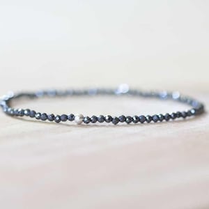 May include: A delicate bracelet featuring small, dark grey beads strung together. A single, silver-coloured bead is interspersed within the design. The bracelet is displayed on a light-coloured wooden surface.