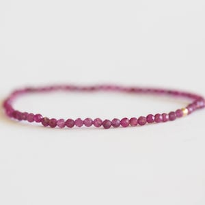 May include: A delicate bracelet featuring small, faceted, round, deep pink gemstones strung together. The bracelet has a simple design with a gold-colored accent. The jewelry is displayed on a white background.