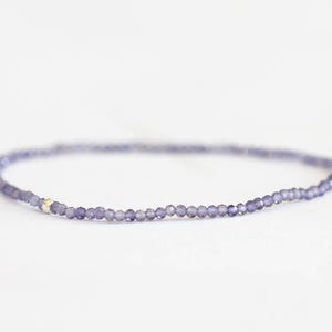 May include: Delicate bracelet featuring small, faceted, lavender-colored beads strung together. A small, silver-colored accent bead is visible. The bracelet is displayed on a white surface, highlighting its subtle elegance. A piece of jewelry.