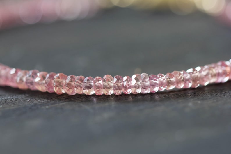 Op de afbeelding: Close-up van een delicate ketting met kleine, gefacetteerde roze edelstenen. De kralen zijn aan elkaar geregen, waardoor een subtiele curve ontstaat. De edelstenen hebben een zachte, doorschijnende uitstraling, wat een elegant effect geeft.