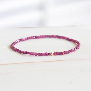 May include: A delicate bracelet composed of small, faceted magenta-colored beads arranged in a circular band. A single, smooth, polished gold-toned spherical bead is positioned as a central accent among the uniform, square-cut magenta beads.