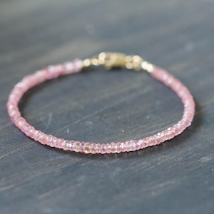 Op de afbeelding: Een delicate roze kralenarmband met een gouden sluiting. De armband is gemaakt van kleine, ronde, lichtroze kralen die aan elkaar zijn geregen. De sluiting heeft een glanzende goudkleur. De armband wordt gepresenteerd op een donkergrijze ondergrond.