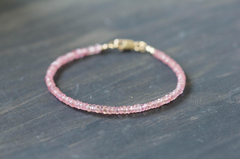 Op de afbeelding: Een delicate roze kralenarmband met een gouden sluiting. De armband is gemaakt van kleine, ronde, lichtroze kralen die aan elkaar zijn geregen. De sluiting heeft een glanzende goudkleur. De armband wordt gepresenteerd op een donkergrijze ondergrond.