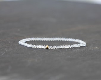 May include: A delicate bracelet composed of small, clear beads. A single gold-colored bead is centered on the bracelet. The bracelet is displayed on a dark gray surface, creating a contrast that highlights the jewelry.