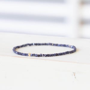 Puede incluir: Una pulsera delicada compuesta por pequeñas cuentas de color azul oscuro de corte cuadrado dispuestas en una banda circular continua. Una sola cuenta esférica lisa y pulida de tono dorado se posiciona como punto focal entre las cuentas azules.