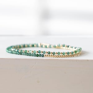 May include: Two delicate bracelets. One is made of small, round, gold beads. The other is made of small, round, green beads. The bracelets are stacked together, creating a layered look. The background is a soft, neutral color.