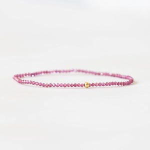 May include: A fine bracelet composed of small, pink, faceted beads. A single gold-coloured bead is positioned centrally. The jewellery is presented against a white backdrop.