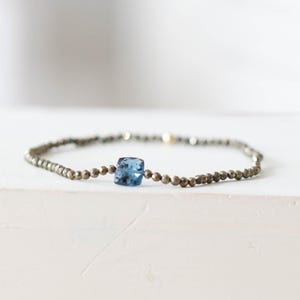 Könnte beinhalten: Ein filigranes Armband mit kleinen, facettierten, metallischen Perlen und einem zentralen, quadratisch geschliffenen, hellblauen Edelstein. Das Armband wird auf einer weißen Oberfläche präsentiert und zeigt sein minimalistisches Design und seine elegante Ästhetik.