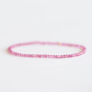 Può includere: Un delicato braccialetto di pietre preziose rosa. Il braccialetto è composto da piccole pietre preziose rosa rotonde e sfaccettate infilate insieme. Il braccialetto è mostrato su uno sfondo bianco.