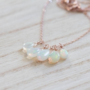 Könnte beinhalten: Eine zarte Halskette mit einer roségoldenen Kette und fünf Opalanhängern. Die Opale haben eine perlmuttartige, irisierende Qualität, und die Halskette wird auf einer hellen Holzoberfläche präsentiert. Schmuck.