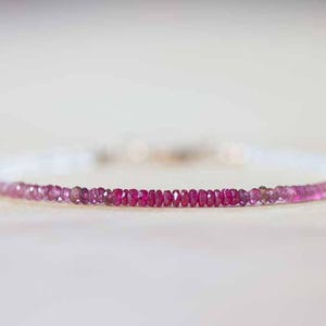 May include: A delicate bracelet featuring small, round, pink gemstones that transition from light to a deep magenta hue. The bracelet is strung on a white band, creating a gradient effect. Jewelry.