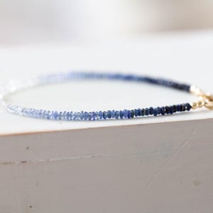 May include: A delicate bracelet featuring a gradient of blue gemstones, transitioning from light to dark. The bracelet is secured with a gold clasp. The gemstones are small and evenly spaced, creating a refined and elegant accessory.
