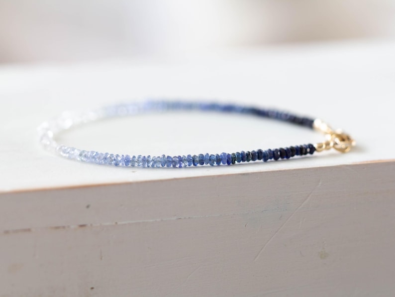 May include: A delicate bracelet featuring a gradient of blue gemstones, transitioning from light to dark. The bracelet is secured with a gold clasp. The gemstones are small and evenly spaced, creating a refined and elegant accessory.