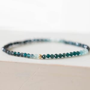 May include: A delicate beaded bracelet featuring small, faceted beads in shades of teal, blue, and a touch of gold. The bracelet is strung on a thin, flexible cord, creating a minimalist and elegant design. The beads are evenly spaced, and the overall aesthetic is refined.