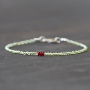 May include: Delicate bracelet featuring small, light green peridot beads with a cluster of red beads at the center. The bracelet has a silver clasp and is displayed on a dark gray surface.
