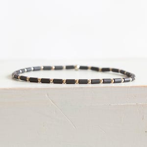 以下が含まれることがあります： マットブラックの円筒形ビーズと、小さな光沢のあるゴールドトーンの球形ビーズが交互に並ぶパターンが特徴のビーズブレスレット。ブレスレットは円形に配置されており、ジュエリー全体を通して一貫した交互のデザインが施されています。