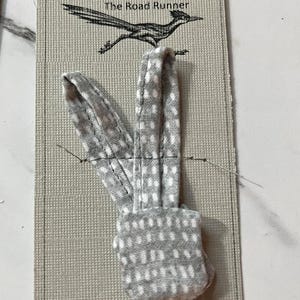 May include: A gray and white polka dot fabric accessory with two upright loops attached to a beige rectangular display card. The card features the text Joe & Jill's ADVENTURE VAN The Road Runner and a black line drawing of a roadrunner bird.
