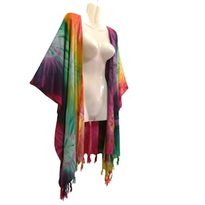 May include: A colourful tie-dye kimono in shades of pink, orange, yellow, green, and purple. The open-front design has wide sleeves and fringed edges. The garment is displayed on a white mannequin, highlighting its flowing silhouette and vibrant colours.