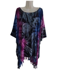 May include: A loose-fitting poncho featuring a vibrant abstract print in shades of deep purple, bright magenta, and black. The garment has a wide, rounded neckline, short sleeves, and a hemline finished with decorative fringe detailing.