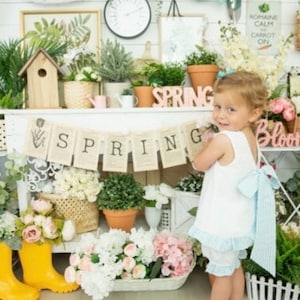 May include: A young girl wearing a white dress with a blue and white striped bow stands in front of a shelf decorated with spring-themed items. The shelf is decorated with flowers, plants, and a banner that reads "SPRING".