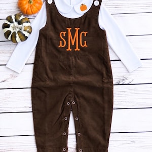 May include: A dark brown corduroy overall featuring a central orange embroidered monogram with the letters SMG, paired with a white long-sleeve shirt that has a Peter Pan collar, a small orange pumpkin embroidery, and white buttons at the shoulders.