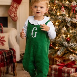 May include: A long-sleeve white shirt with a rounded collar worn under a solid green overall featuring a white embroidered letter G on the chest. The overall has a button-down front closure and is paired with simple white shoes for a complete outfit.
