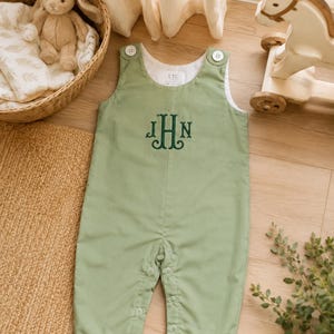 May include: A sage green sleeveless romper featuring a dark green embroidered monogram with the letters JHN on the chest. The garment has white buttons at the shoulders and a snap-button closure along the inner legs, with an ETC brand label inside.