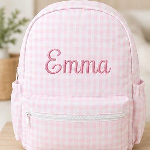 May include: A light pink and white gingham patterned backpack featuring the name Emma embroidered in pink script lettering on the front. The backpack includes a front zippered pocket, two side pockets, and white zipper hardware.