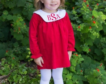May include: A young child wearing a red dress with a white collar and the embroidered initials "BSA". The dress has long sleeves and is paired with white tights and sneakers. A large white bow adorns the child's hair. The background features green foliage.