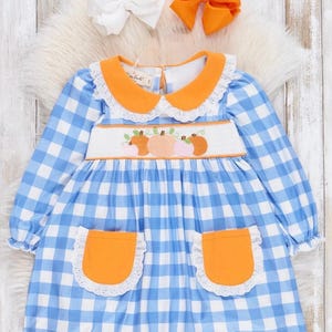 May include: A blue and white gingham dress with orange trim and pockets. The dress has a white lace collar and embroidered pumpkins on the bodice.