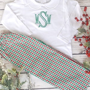 May include: A white long-sleeved shirt with a Peter Pan collar and green embroidered initials, paired with red and green plaid pants. The outfit is displayed on a light wood surface with holiday greenery.
