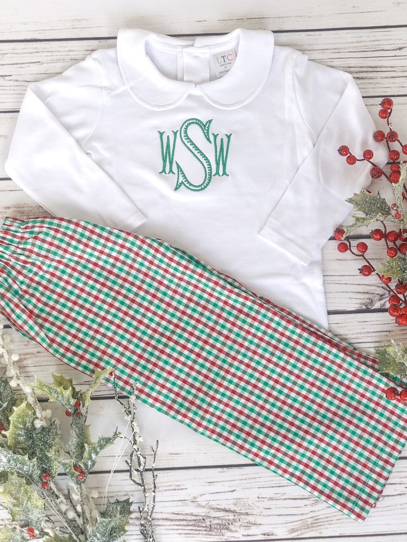 May include: A white long-sleeved shirt with a Peter Pan collar and green embroidered initials, paired with red and green plaid pants. The outfit is displayed on a light wood surface with holiday greenery.