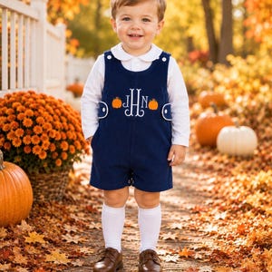May include: A navy blue corduroy overall short set featuring a white long-sleeve collared shirt, white knee-high socks, and brown leather shoes. The overalls are embroidered with a white monogram and two orange pumpkins on the chest area.