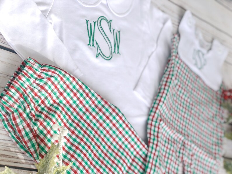 May include: White long-sleeved shirt and matching pants with a red and green plaid pattern. The shirt has a Peter Pan collar and a green embroidered monogram. The outfit is designed for children and is suitable for the holiday season.