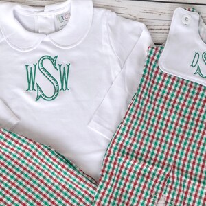 May include: White long-sleeved shirt and matching pants with a green monogram. The pants and romper feature a red and green plaid pattern. A white bib with a green monogram is also included.