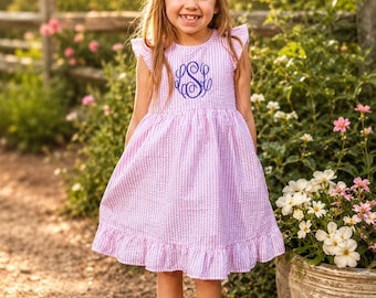 May include: A light purple and white gingham dress with ruffled sleeves and a ruffled hem. The dress features a dark purple embroidered monogram on the chest. A large purple hair bow is worn on the head, and silver metallic sandals are worn.