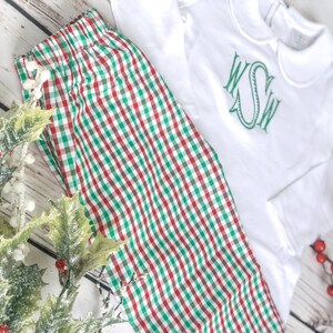May include: White long-sleeved shirt with a green monogram and a red, green, and white checkered pajama set. The pajamas are made of a soft fabric. The outfit is displayed with holly and red berries, suggesting a holiday theme.