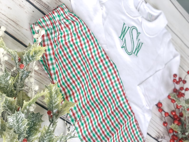 May include: White long-sleeved shirt with a green monogram and a red, green, and white checkered pajama set. The pajamas are made of a soft fabric. The outfit is displayed with holly and red berries, suggesting a holiday theme.