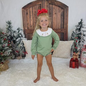 May include: A young child wearing a sage green romper with a white collar and trim. The collar features embroidered reindeer and the letter "F". The child has a large red bow in their hair. The background includes Christmas decorations and a wooden door.