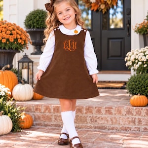 May include: A brown corduroy jumper dress featuring a white long-sleeve collared shirt underneath. The dress has an orange embroidered monogram on the chest, a small pumpkin accent on the collar, and white buttons at the shoulders.